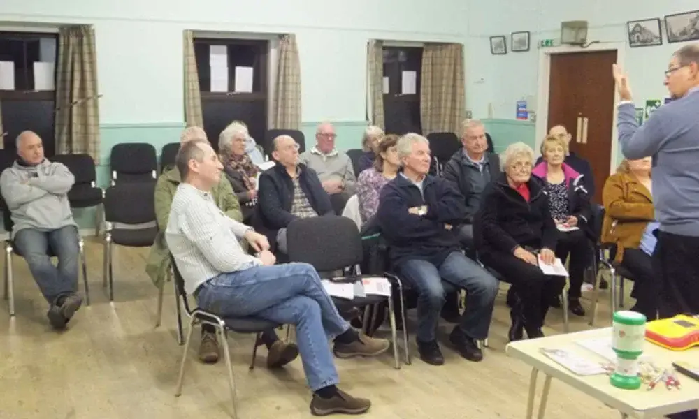 Group of adults seated and listening to a speaker in a community room with wooden floors.