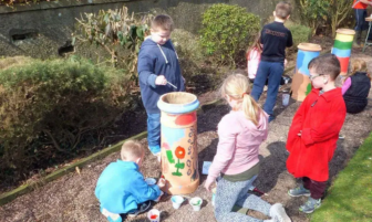 Children painting large outdoor pots with colourful designs along a garden path.